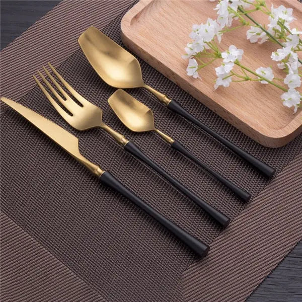 Set of gold cutlery with black handles on a brown placemat with a wooden board and flowers in the background.