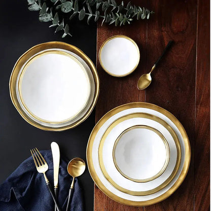 Set of gold-rimmed white plates and silverware on a dark surface with greenery.