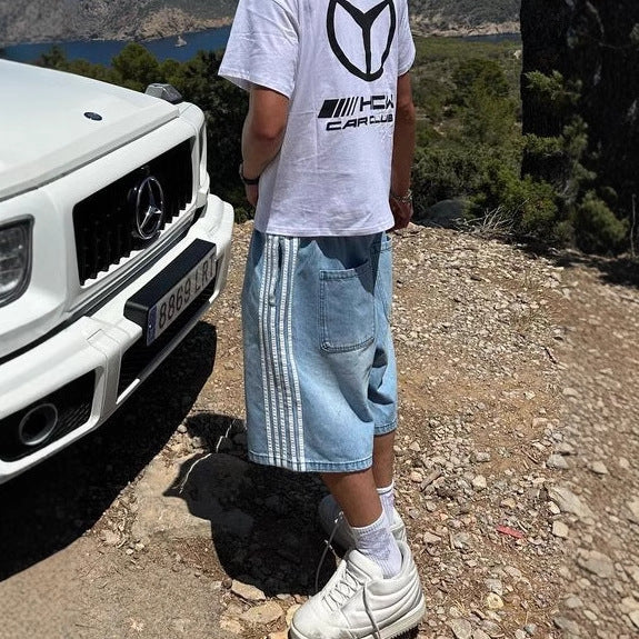 Person wearing a white t-shirt with a logo and blue shorts standing next to a white Mercedes-Benz car on a scenic road.
