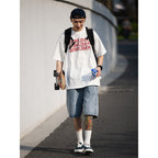 Person wearing a white t-shirt with text, denim shorts, and a black cap, walking outdoors.