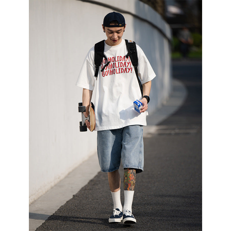 Person wearing a white t-shirt with text, denim shorts, and a black cap, walking outdoors.